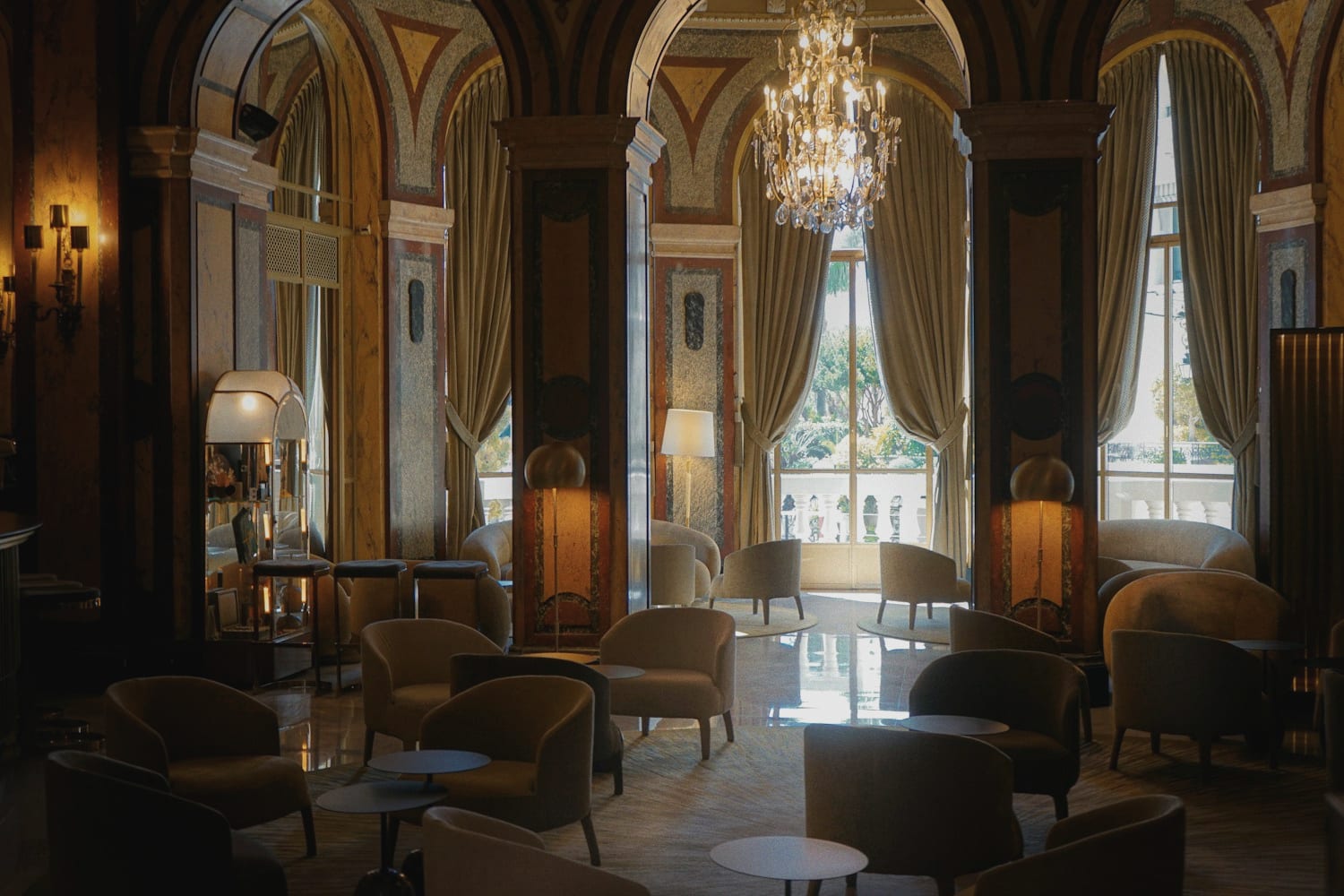 The lounge of a grand hotel: marble columns under painted vaulted arches, a crystal chandelier, and armchairs drawn up to tall curtained windows that open onto a balustraded terrace.