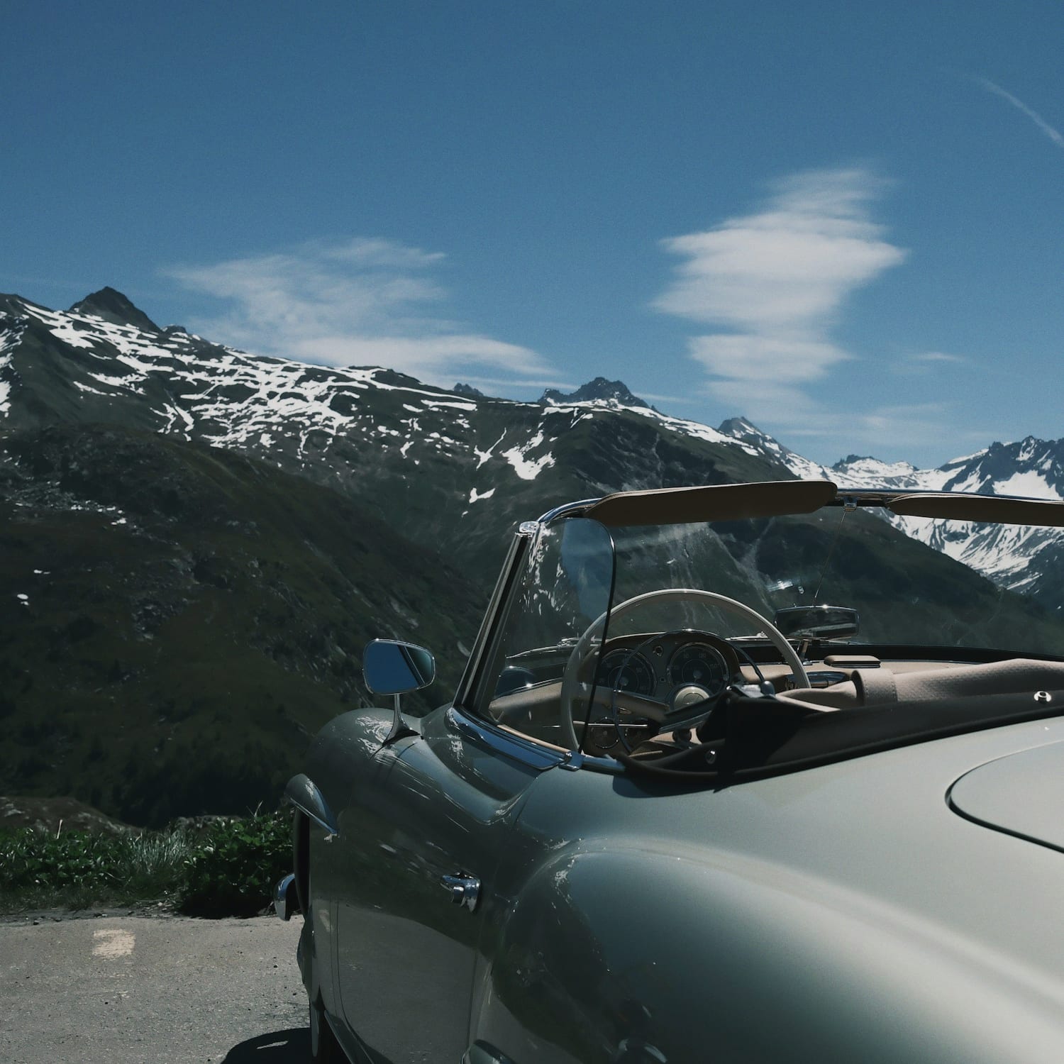 A vintage Mercedes 190SL roadster stopped at an alpine pass, its ivory steering wheel and chrome cockpit open to snow-streaked peaks under a deep blue sky.