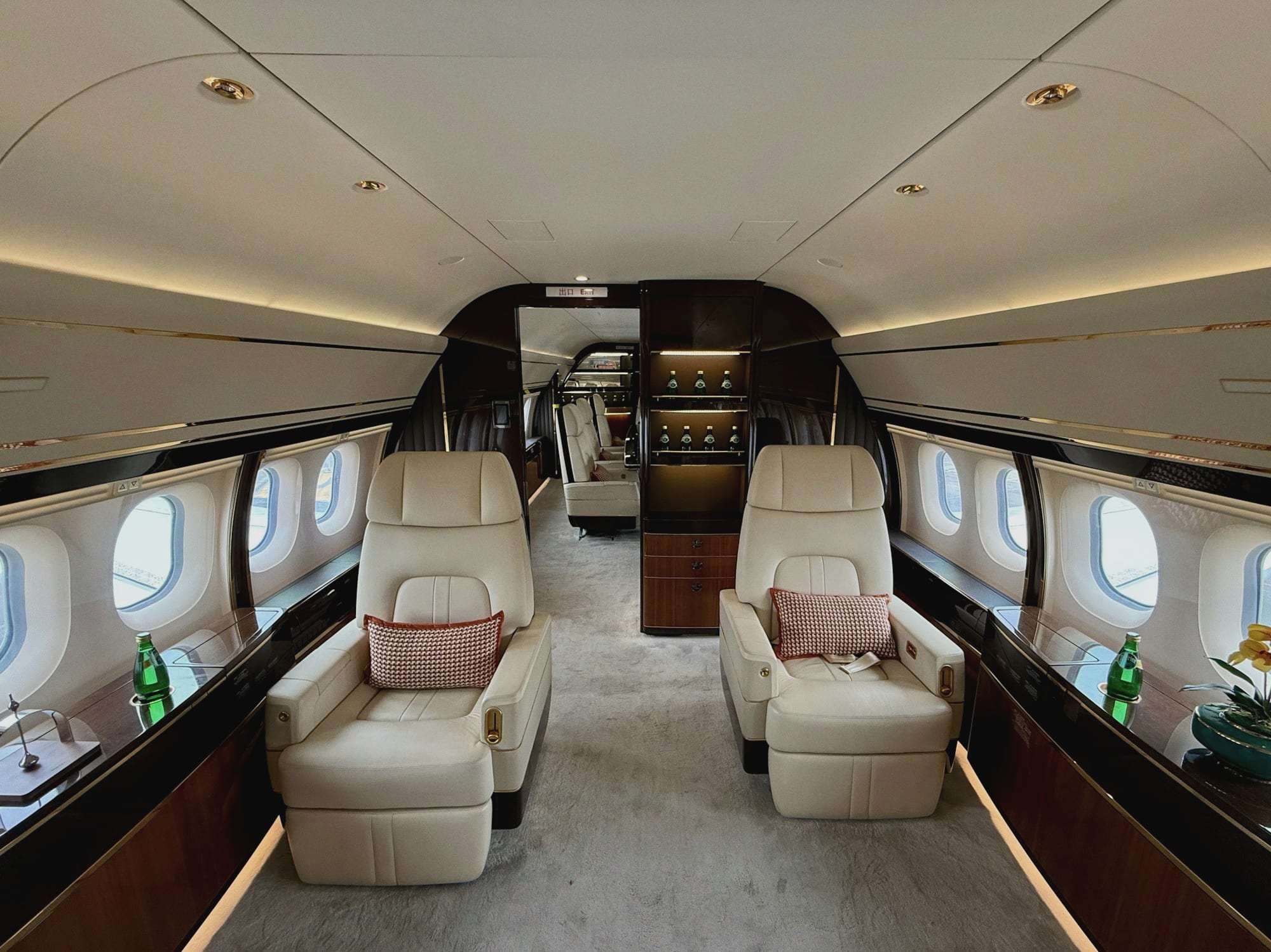 The cabin of a private jet: cream leather club seats facing each other across the aisle, walnut veneer cabinets and a bar, lit by a warm line of cove light along the ceiling.